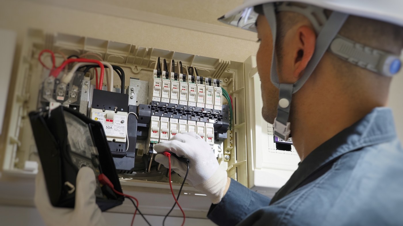 ヘルメットをかぶった職人が、扉を開けた分電盤を測定器で調べている